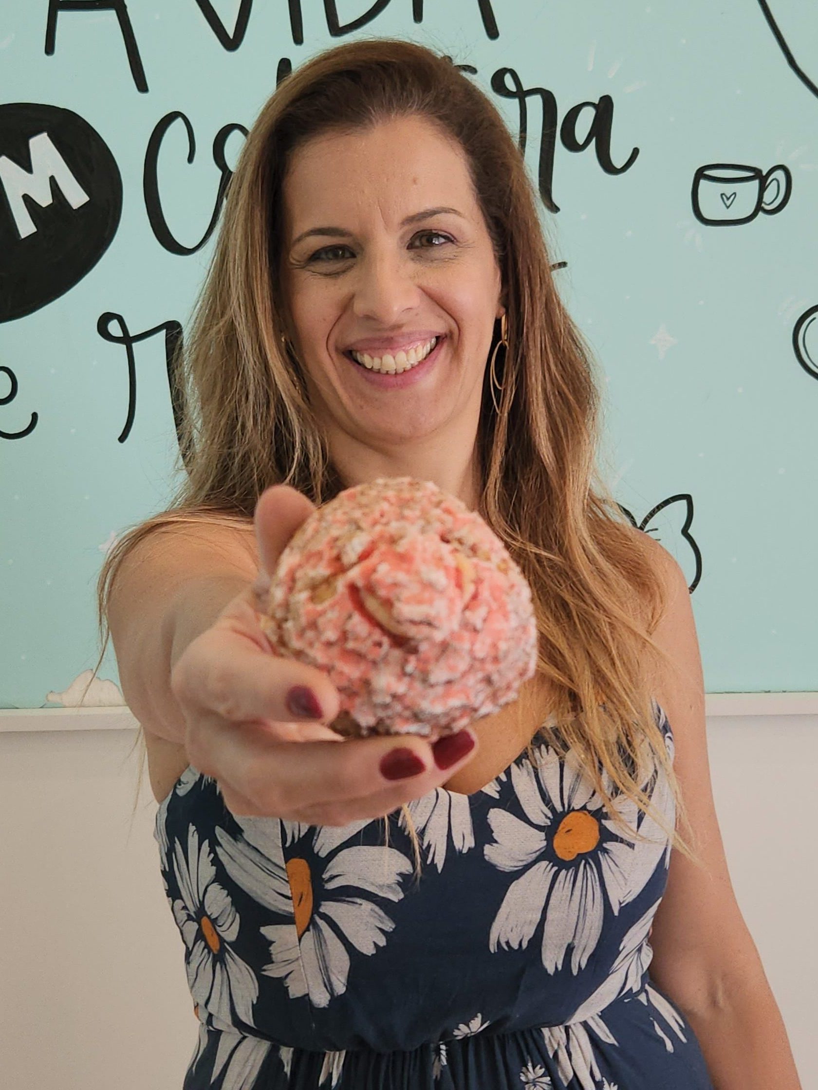 Lu Veras sorrindo enquanto oferece uma choux cream coberta com crocante colorido, em frente ao mural da confeitaria As Veras.